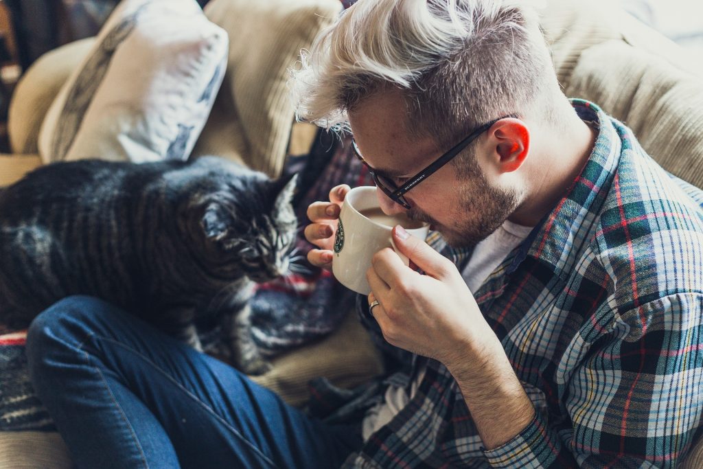 man sipping a cup on coffee while sitting on a couch with a black cat