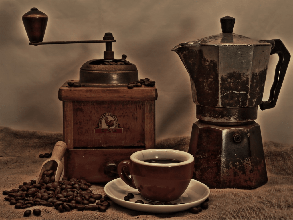 old fashioned coffee grinder and coffee pot with a cup and saucer and coffee beans in the foreground
