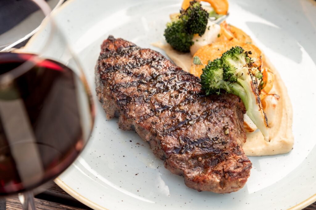 New York strip steak, alongside broccoli and a creamy mash on a white plate and a glass of red wine