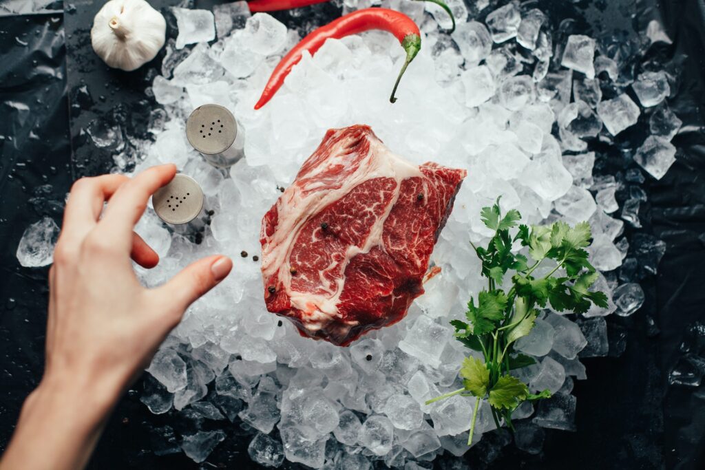 uncooked porterhouse steak sitting on a bed of ice - a woman's hand is reaching for salt and pepper shakers